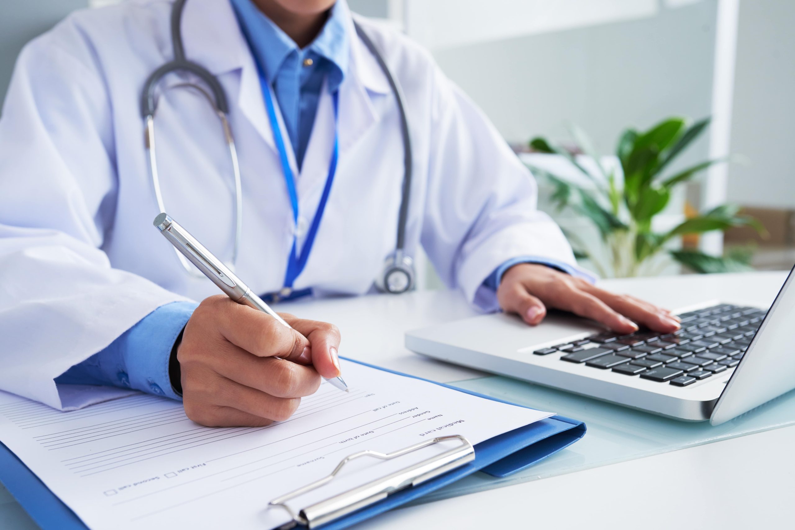 hands-unrecognizable-female-doctor-writing-form-typing-laptop-keyboard-min