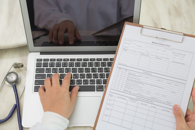 A doctor using a laptop and reviewing a clipboard with patient information on a desk in a medical office.